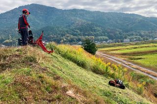 田園風景の草が茂った斜面で稼働するロボット芝刈り機を観察する作業用具を着た人。